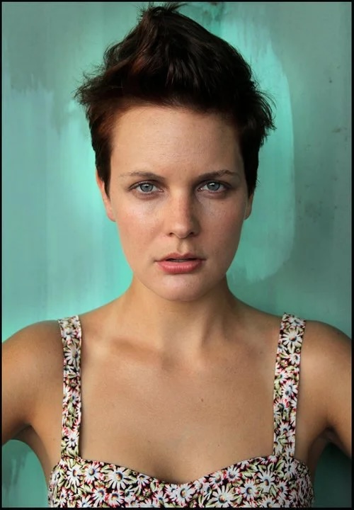 Person with short brown hair posing against a teal background, wearing a floral dress.