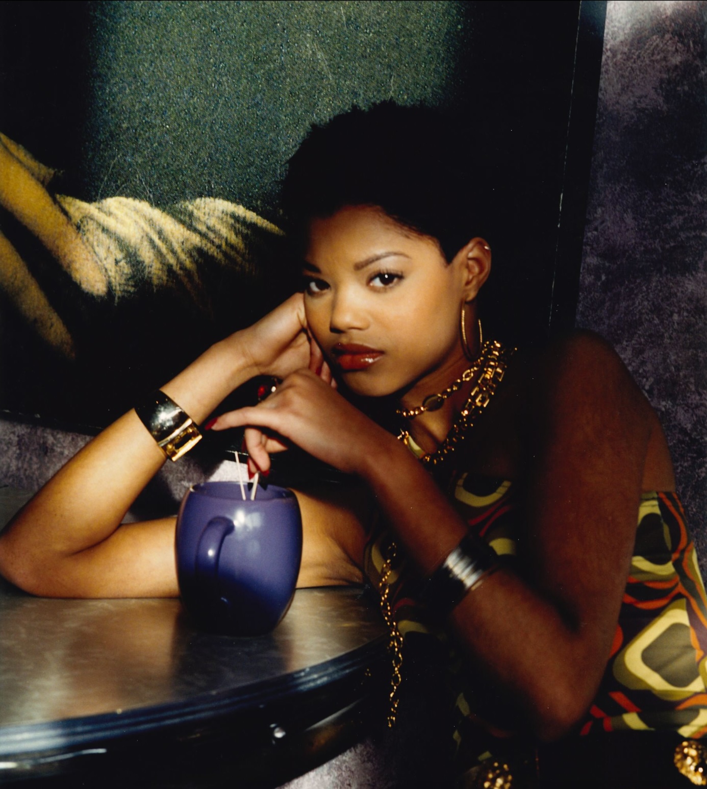 Woman with short hair sitting at a table, holding a drink in a purple mug, wearing bold jewelry and a patterned top.