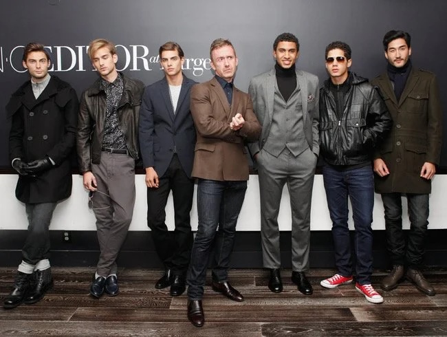 A group of men in fashion-forward outfits, posing against a dark-colored wall.